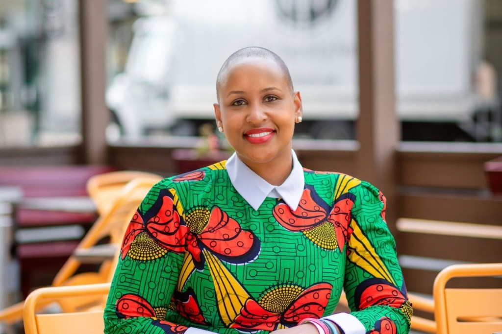 A woman with a shaved head smiling, wearing a vibrant green sweater adorned with colorful patterns, sitting outdoors at a cafe.