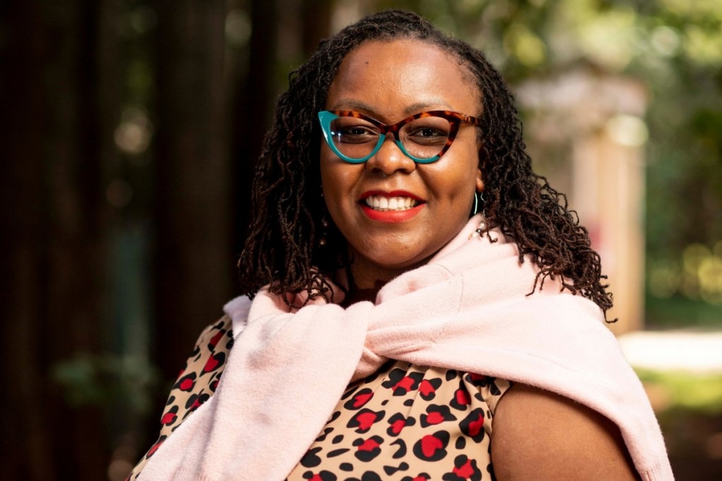 A woman with dark, curly hair wearing glasses and a patterned dress, smiling while wrapped in a light pink scarf, standing outdoors among trees.